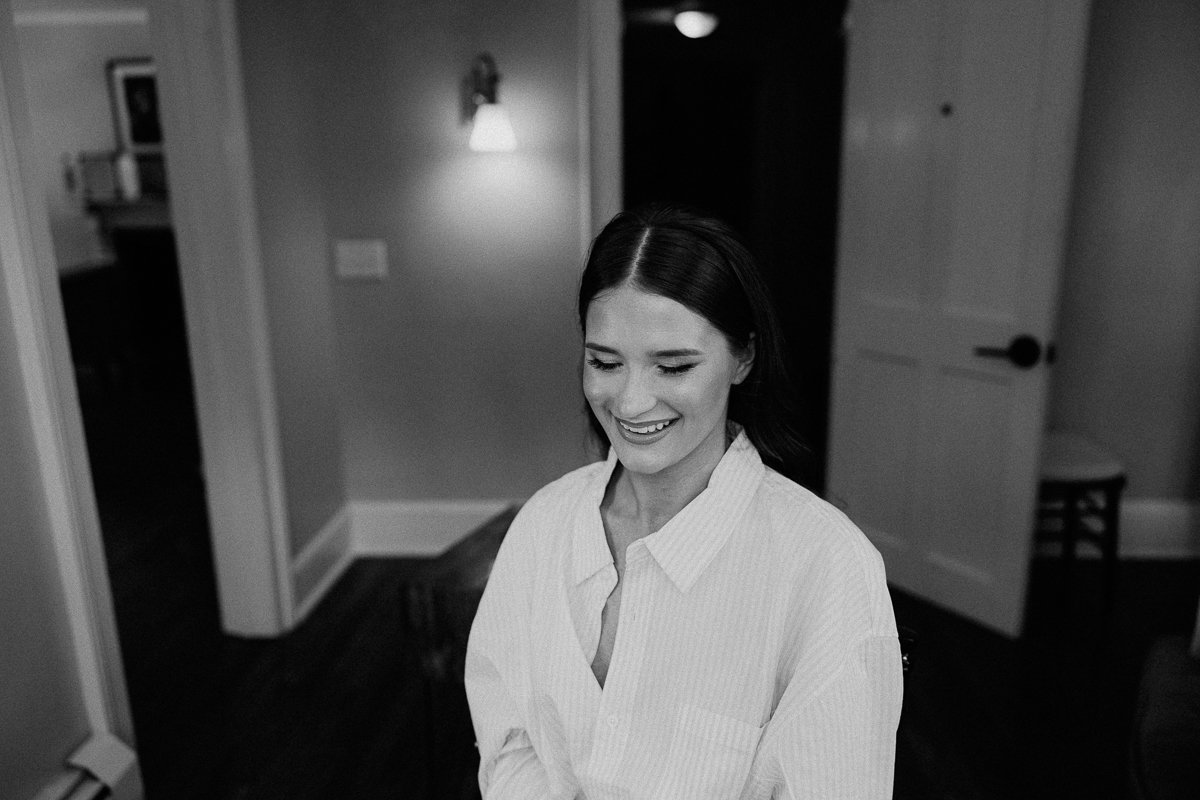 A woman with straight, dark hair and a light-colored shirt sits indoors, smiling and looking down. The black and white photo, softly lit with a door and wall lamp in the background, captures a quiet moment before a Seven Hills Inn wedding in Lenox MA.