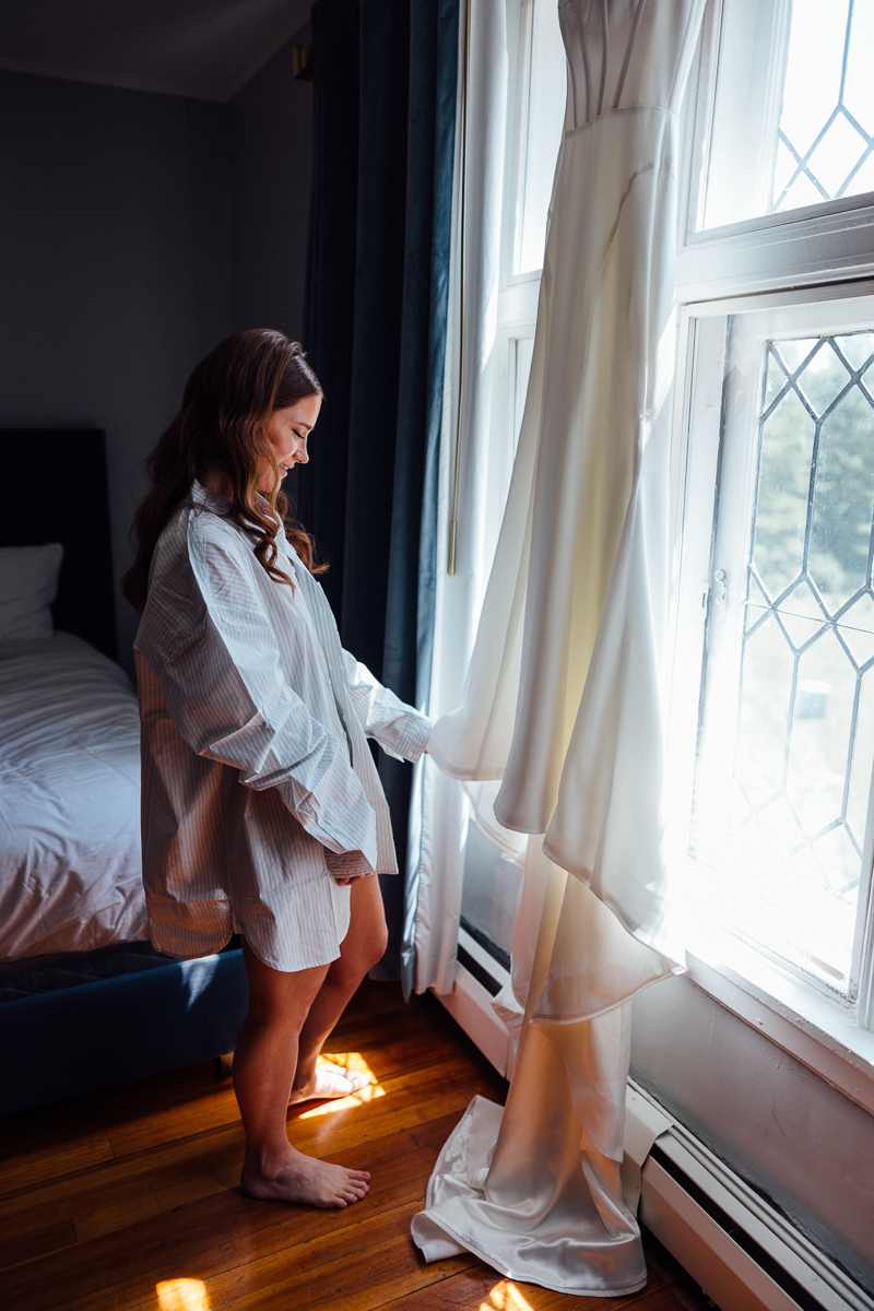 A woman in an oversized shirt stands barefoot by a window at Seven Hills Inn in Lenox MA, gently touching a white satin wedding dress hanging beside her. Sunlight streams in, illuminating the dress and the wooden floor.