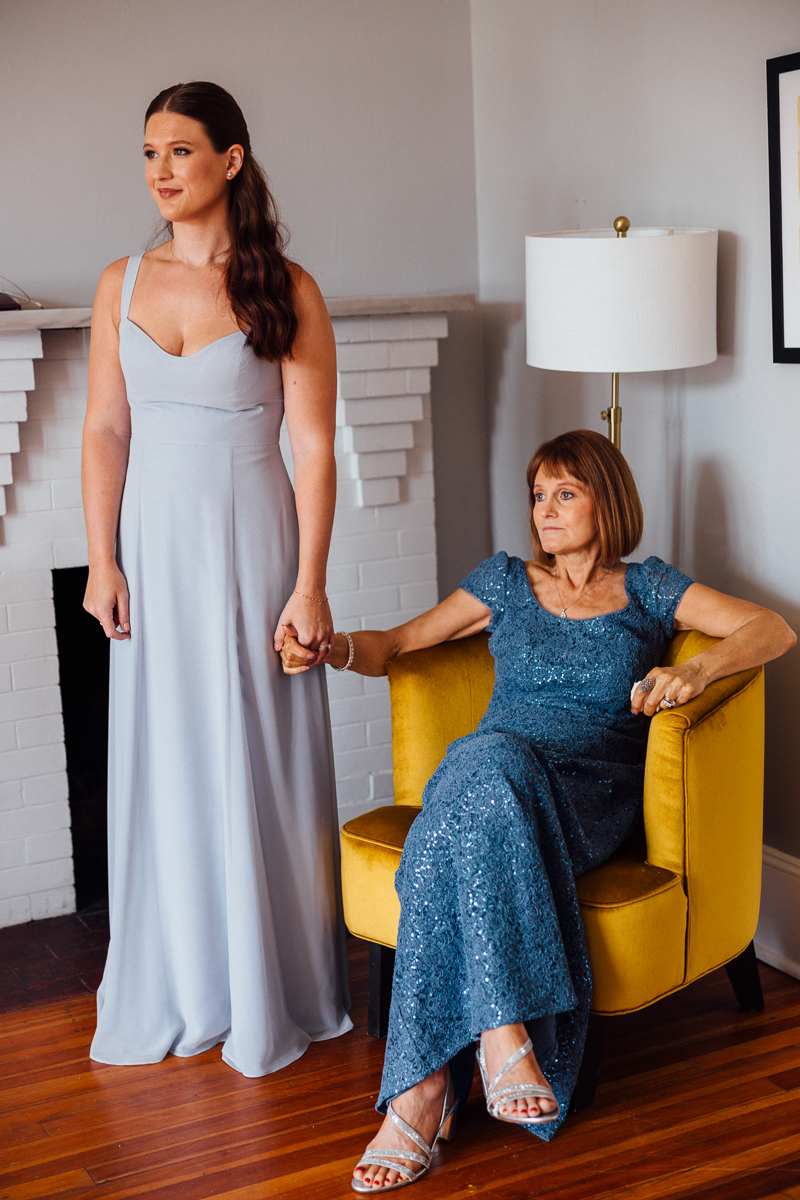 At Seven Hills Inn in Lenox, MA, a woman in a light blue gown stands holding hands with an older woman in a blue sequin gown seated by a yellow chair. They share a heartfelt moment indoors near a white fireplace and floor lamp.