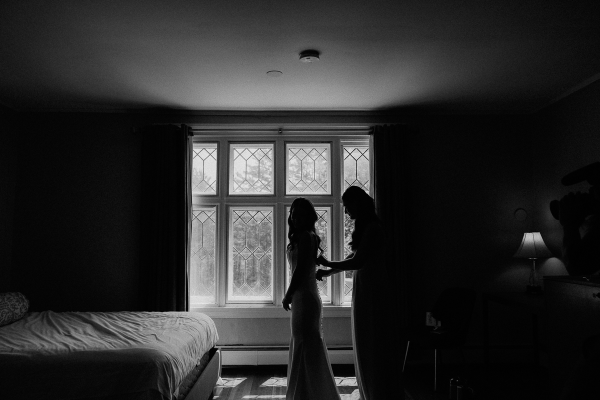 A silhouetted bride stands near a large window at Seven Hills Inn in Lenox, MA, as another woman helps her with her dress in a softly lit bedroom, with light streaming in from outside during this intimate wedding moment.