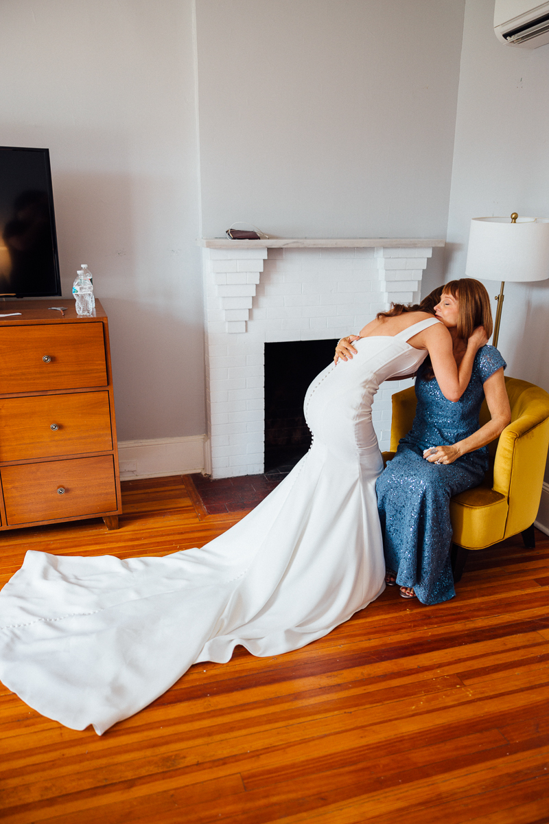 At a Seven Hills Inn Wedding in Lenox MA, a bride in a white gown bends to hug a woman in a blue dress on a yellow chair, sharing an emotional moment in a cozy room with hardwood floors and a white fireplace.