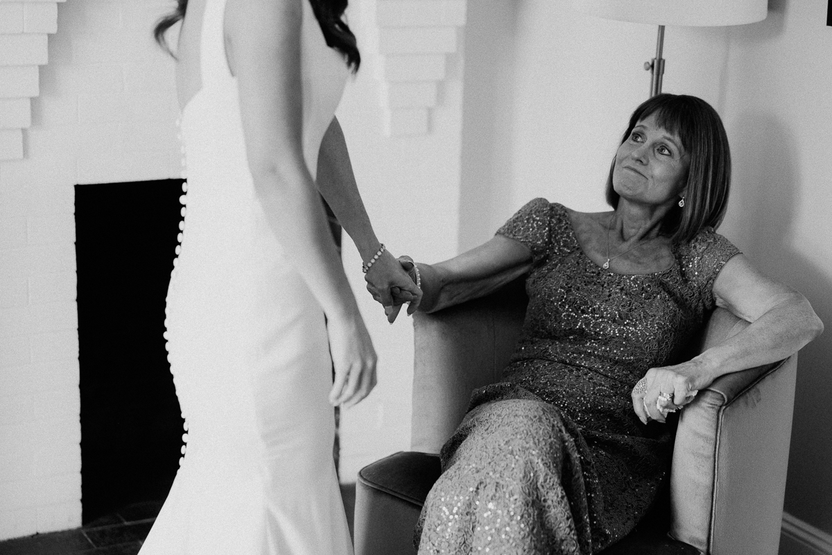 At the Seven Hills Inn in Lenox, MA, a woman in a sparkly dress sits smiling and holding hands with a bride in a wedding dress. They share an emotional, intimate wedding moment indoors.