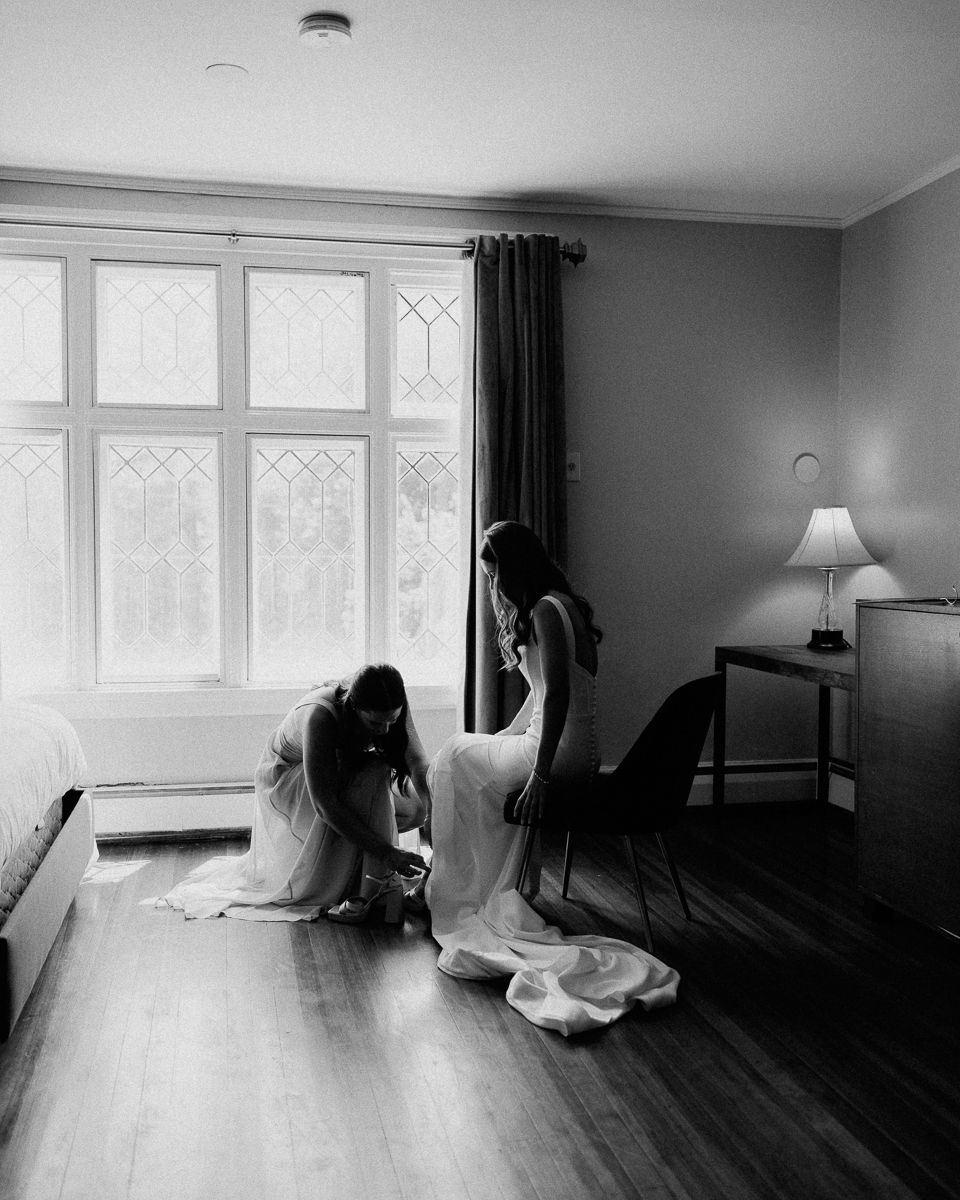 Two women in dresses are in a softly lit room at the Seven Hills Inn in Lenox, MA; one sits while the other kneels to adjust her dress train. Sunlight filters through large windows, creating a calm, intimate atmosphere.