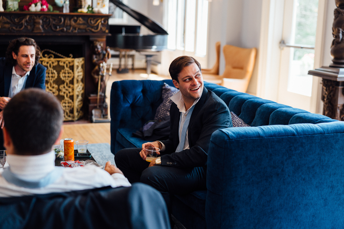 A man in a suit smiles while holding a drink, seated on a blue velvet sofa in a stylish living room at a Seven Hills Inn Wedding in Lenox, MA. Two other men in suits are present, one facing away and one by the decorative fireplace.