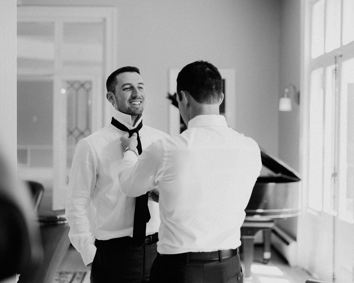 Two men in dress shirts and ties stand indoors at a Seven Hills Inn Wedding in Lenox, MA; one helps the other adjust his tie as they smile. Soft natural light fills the room, and a grand piano is visible in the black-and-white photo.