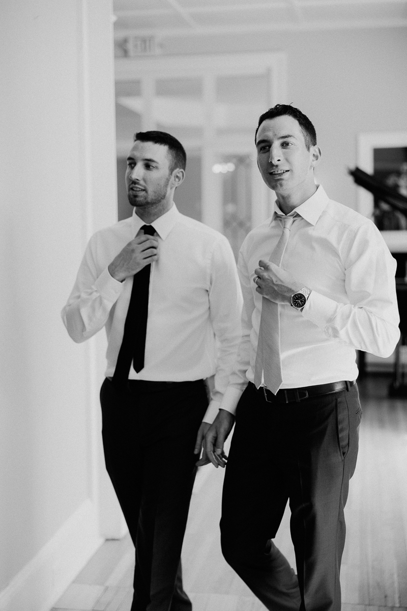 Two men in dress shirts and ties walk indoors, adjusting their ties. This black and white photo, likely taken before a Seven Hills Inn wedding in Lenox MA, captures them preparing amid hallway details and a piano.
