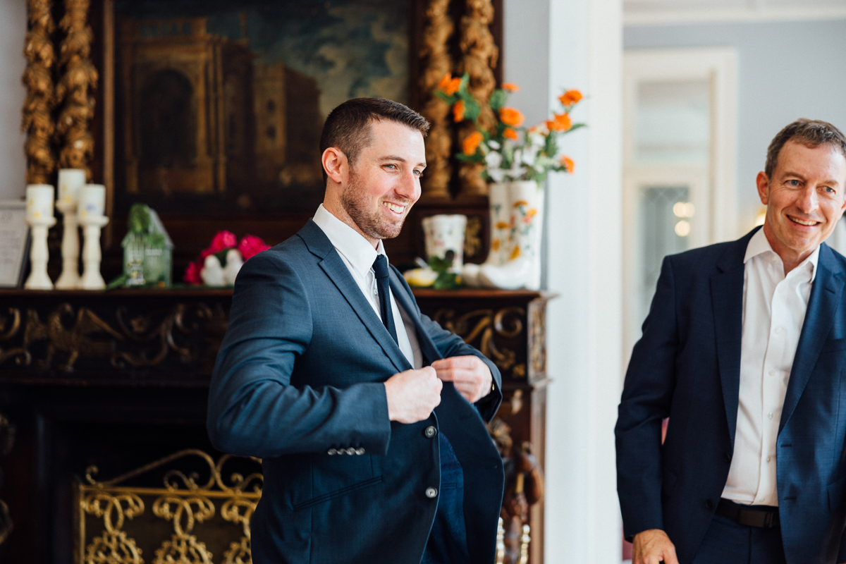A man in a suit smiles while buttoning his jacket, standing indoors near an ornate fireplace with flowers and a painting at a Seven Hills Inn wedding in Lenox, MA. Another man in a suit stands nearby, also smiling.