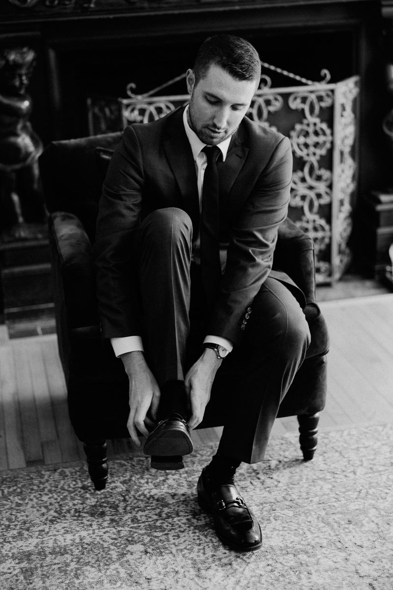 A man in a suit sits in an armchair, looking down as he adjusts one of his dress shoes. The formal setting, with its ornate fireplace and patterned screen, hints at an elegant Seven Hills Inn Wedding in Lenox MA. The photo is in black and white.