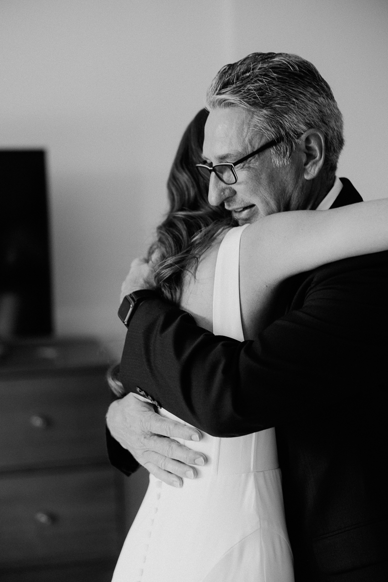 A black and white photo of a man in glasses and a suit warmly hugging a woman in a white dress at their Seven Hills Inn wedding in Lenox, MA, capturing an emotional, intimate moment.