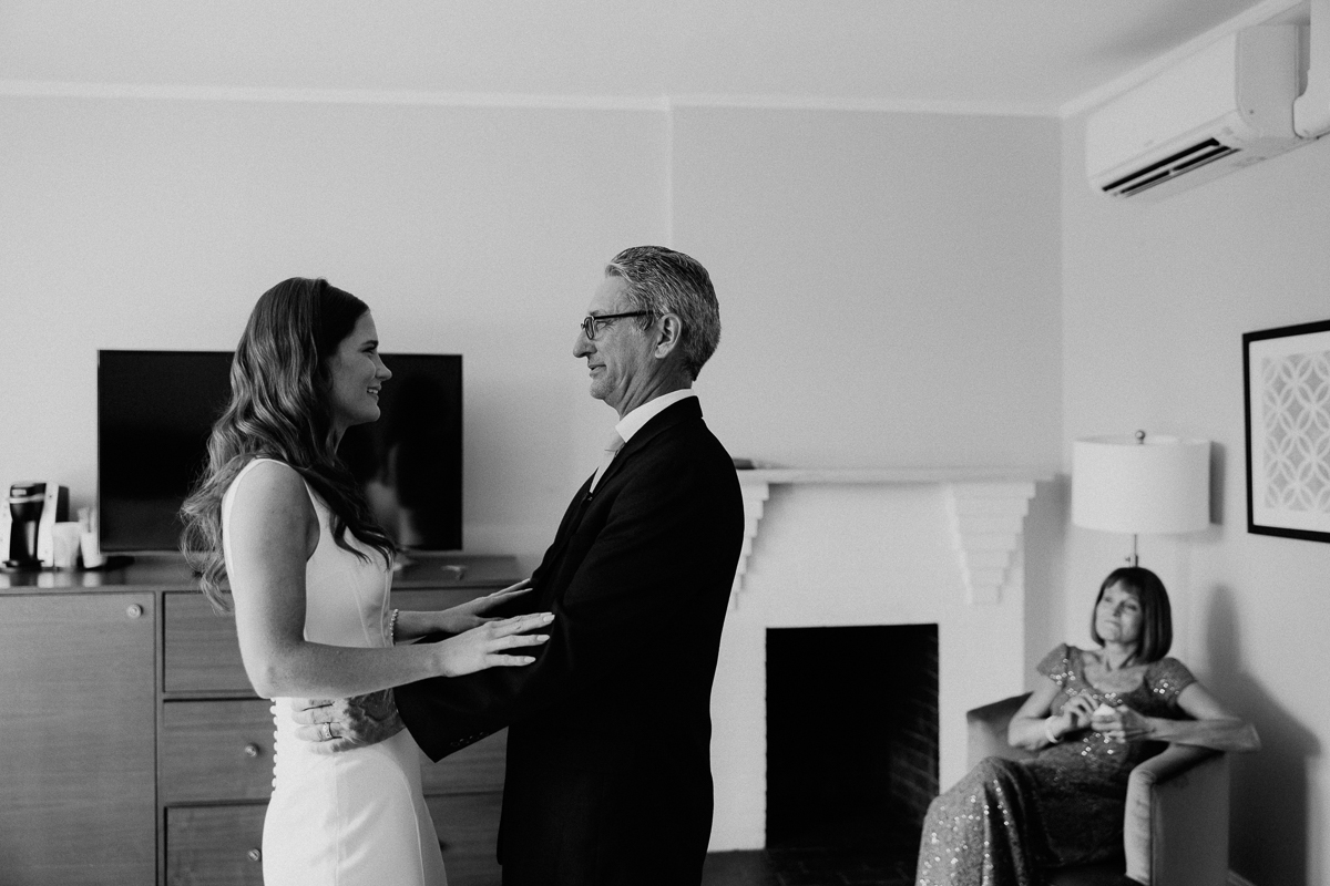 A bride and an older man, both smiling, share a touching moment while holding hands in a room at the Seven Hills Inn in Lenox, MA. A woman in a dress sits on a chair in the background, watching them warmly. The wedding scene is in black and white.