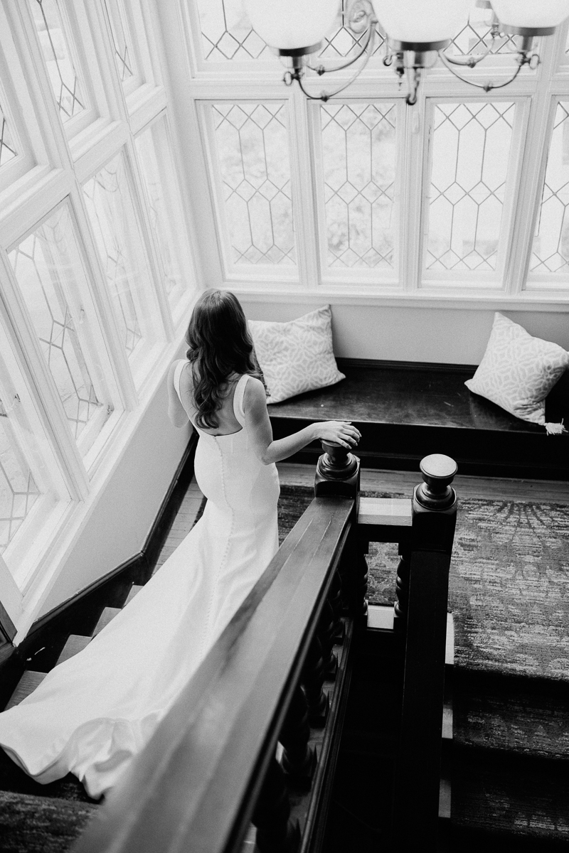 A bride in a white gown stands at the top of a staircase, looking out sunlit patterned-glass windows. This elegant and peaceful moment at Seven Hills Inn in Lenox MA features two pillows on a nearby window seat.