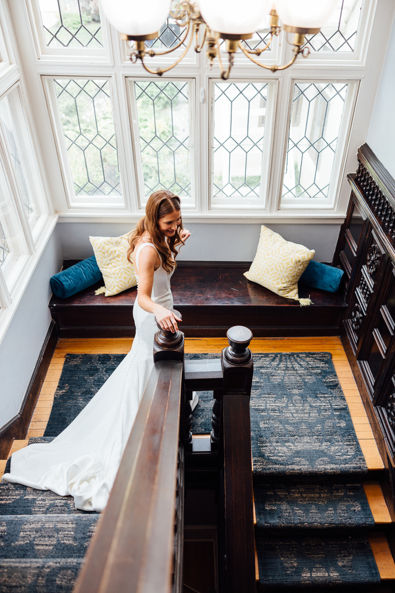A bride in a white wedding dress stands on a wooden staircase at Seven Hills Inn, Lenox MA, gazing down, with large windows and built-in benches with pillows behind her in a well-lit room.