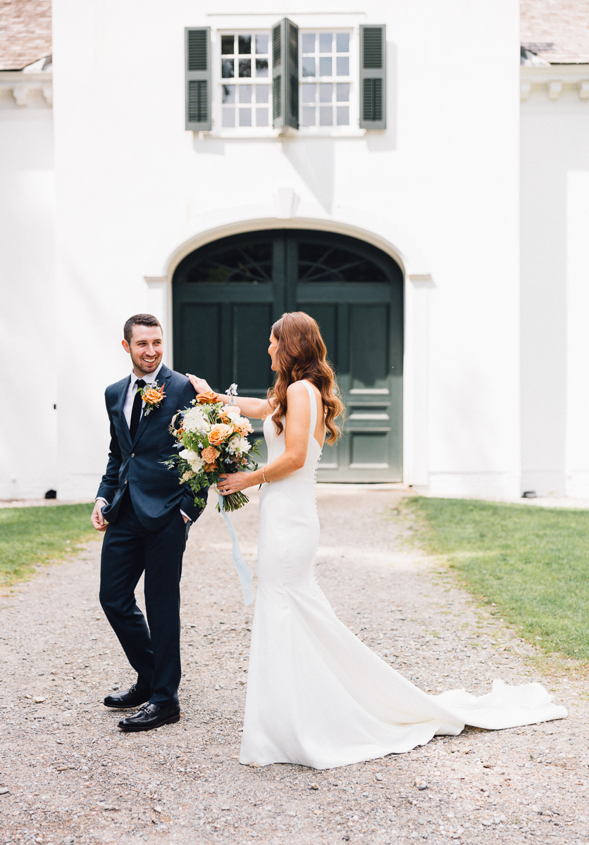 A bride in a white dress holding a bouquet smiles at her groom in a dark suit as they stand outside Seven Hills Inn in Lenox MA, enjoying a joyful wedding moment together by the green doors and open shutters.