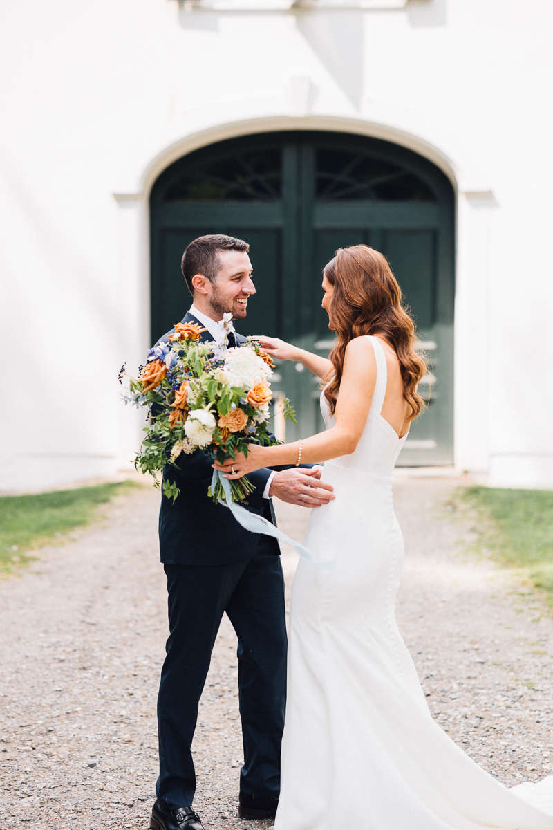 A bride in a white dress and a groom in a dark suit smile at each other outside Seven Hills Inn in Lenox MA, with the bride holding a large bouquet of orange and white flowers before green double doors.