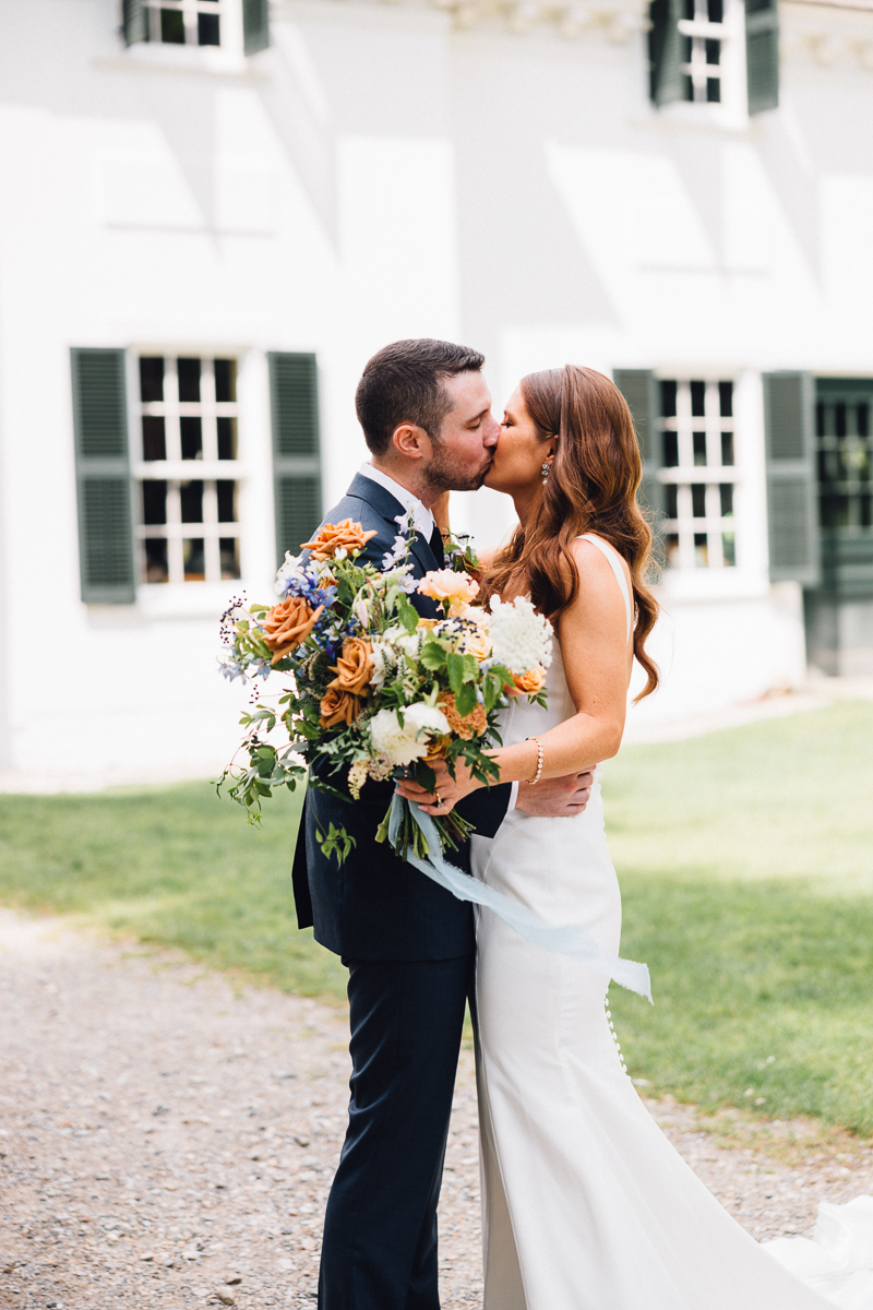 A bride in a white dress and groom in a dark suit kiss outdoors at their Seven Hills Inn wedding, holding a large bouquet of orange, white, and green flowers, with a white house and green shutters in the background in Lenox MA.