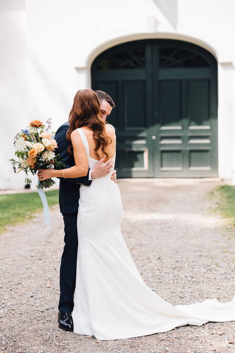 A bride in a white dress holding a bouquet hugs a groom in a dark suit outdoors, standing on a gravel path in front of large dark green double doors at the beautiful Seven Hills Inn, a charming Lenox Massachusetts wedding venue.