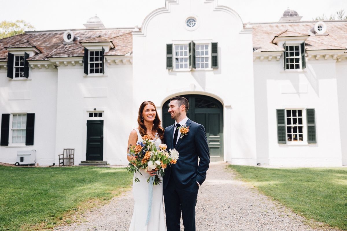 A bride and groom stand smiling outside the elegant Seven Hills Inn, a renowned Lenox Massachusetts Wedding Venue with green shutters and an arched doorway, holding a colorful bouquet on a sunny day.