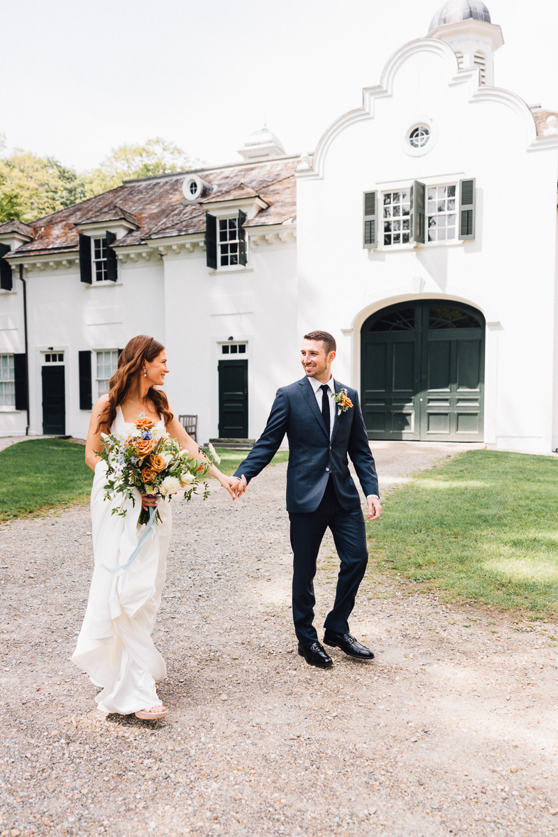 A bride in a white dress holds a bouquet and smiles at her groom as they walk hand-in-hand outside the historic Seven Hills Inn, a stunning Lenox Massachusetts wedding venue with grand green doors and shuttered windows.