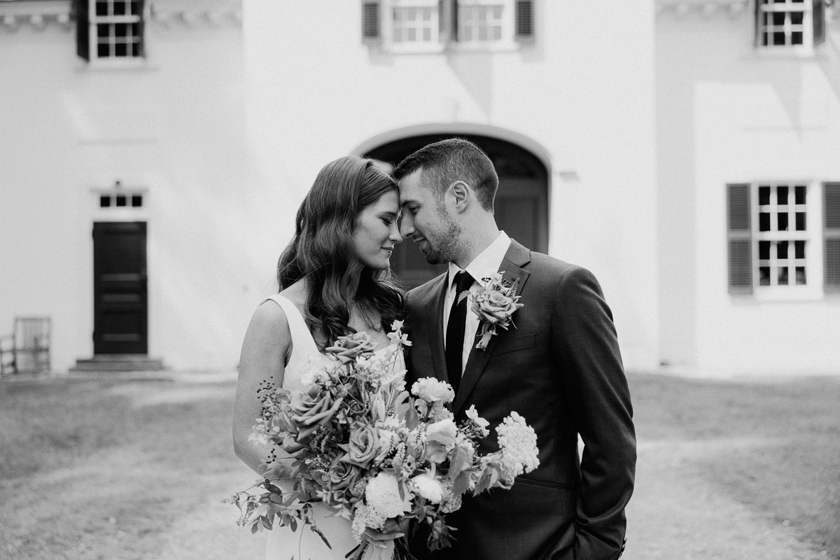 A bride and groom stand close together, smiling softly and holding a bouquet in front of the elegant Seven Hills Inn, a beloved Lenox Massachusetts wedding venue. The black and white image creates a timeless, romantic atmosphere.