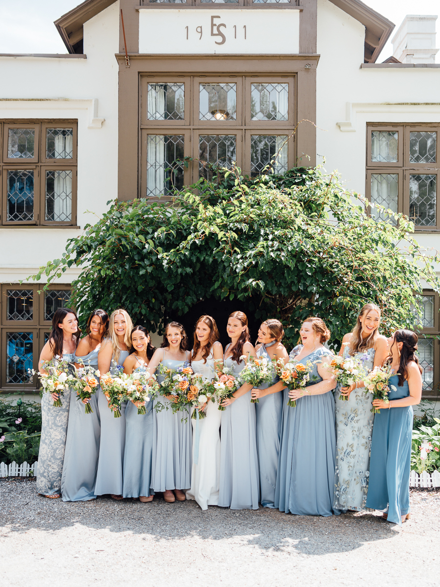 A bride stands smiling with her bridesmaids, all holding flower bouquets and wearing light blue, grey, and patterned dresses, in front of a leafy green bush and the historic Seven Hills Inn—a beautiful Lenox Massachusetts Wedding Venue.