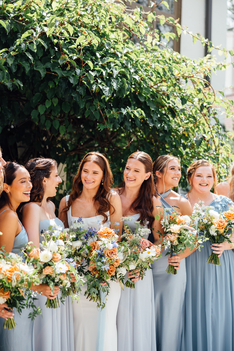 A group of smiling bridesmaids in light blue dresses stand together outdoors at Seven Hills Inn, a beautiful Lenox Massachusetts Wedding Venue, holding colorful floral bouquets with greenery and sunlight in the background.