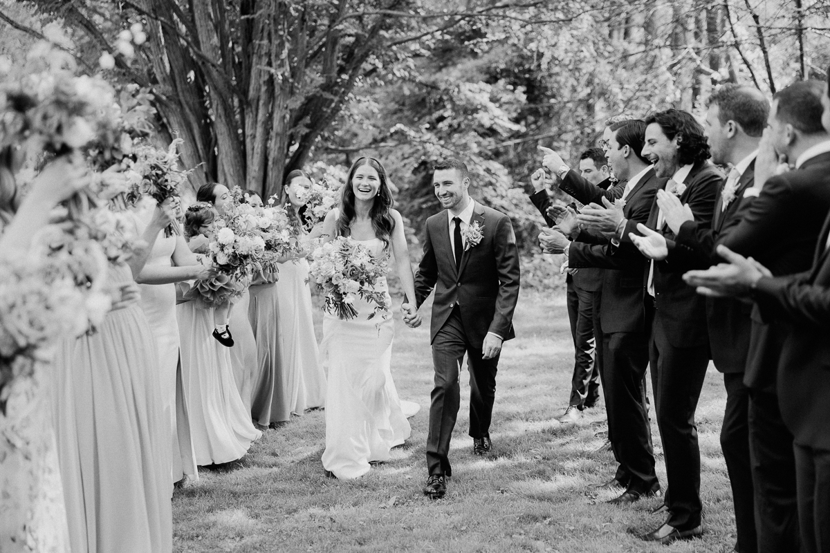A bride and groom walk hand in hand outdoors at the Lenox Wedding, smiling as their wedding party stands on either side, cheering and holding bouquets; the scene is joyful and surrounded by trees at this Lenox Massachusetts Wedding Venue.