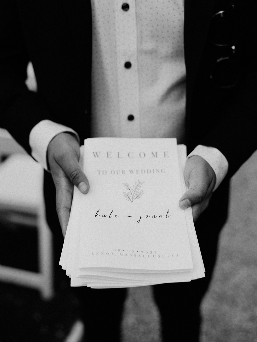 A person in a suit holds a stack of wedding programs that read, Welcome to our wedding, Kate + Jonah, 09.03.2023, Lenox, Massachusetts, in elegant script—a timeless black and white moment from a classic Lenox Wedding at Seven Hills Inn.