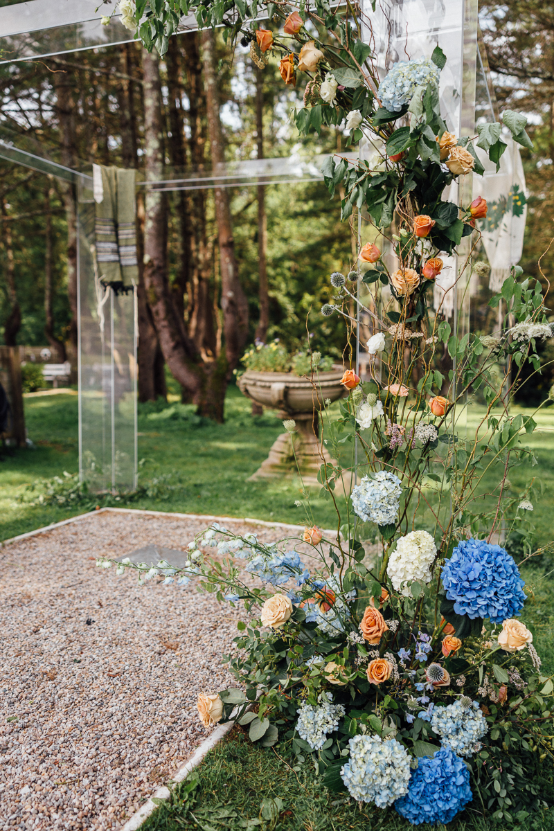 A floral wedding arch at Seven Hills Inn, adorned with orange roses and blue hydrangeas, stands on a gravel path. Green grass, trees, and a blurred stone birdbath create a picturesque Lenox Massachusetts Wedding Venue backdrop.