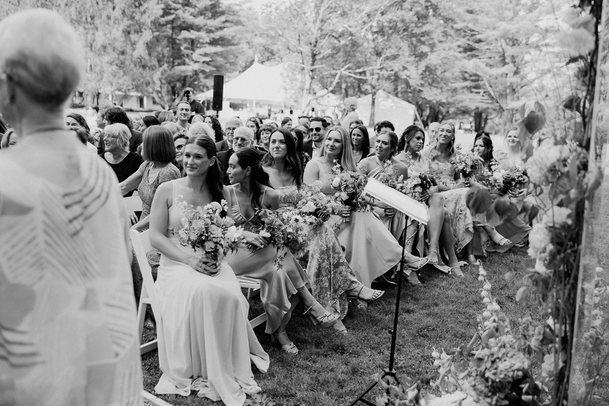 A group of bridesmaids in light dresses sit in rows holding bouquets at an outdoor Lenox Wedding ceremony, surrounded by guests, trees, and flowers on a grassy lawn.