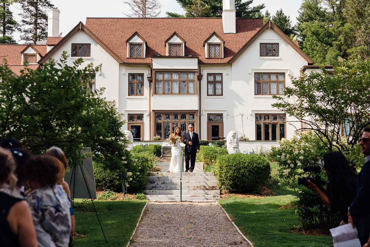 A bride in a white dress walks down stone steps with a man in a suit, surrounded by greenery at Seven Hills Inn, a stunning Lenox Massachusetts Wedding Venue, as guests watch and an elegant house stands in the background.