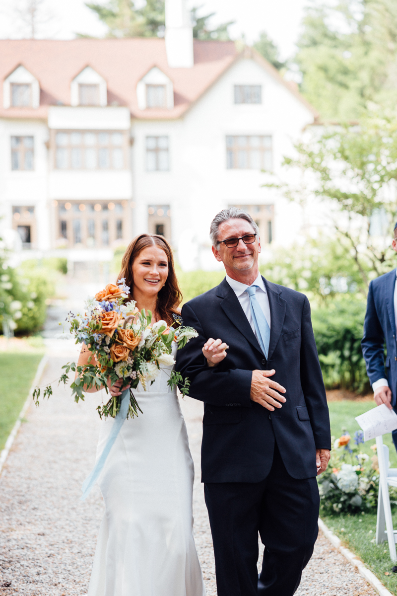 A smiling bride in a white dress holding a bouquet walks arm-in-arm with a man in a dark suit down an outdoor path at Seven Hills Inn, a picturesque Lenox Massachusetts wedding venue framed by greenery and a grand white house.