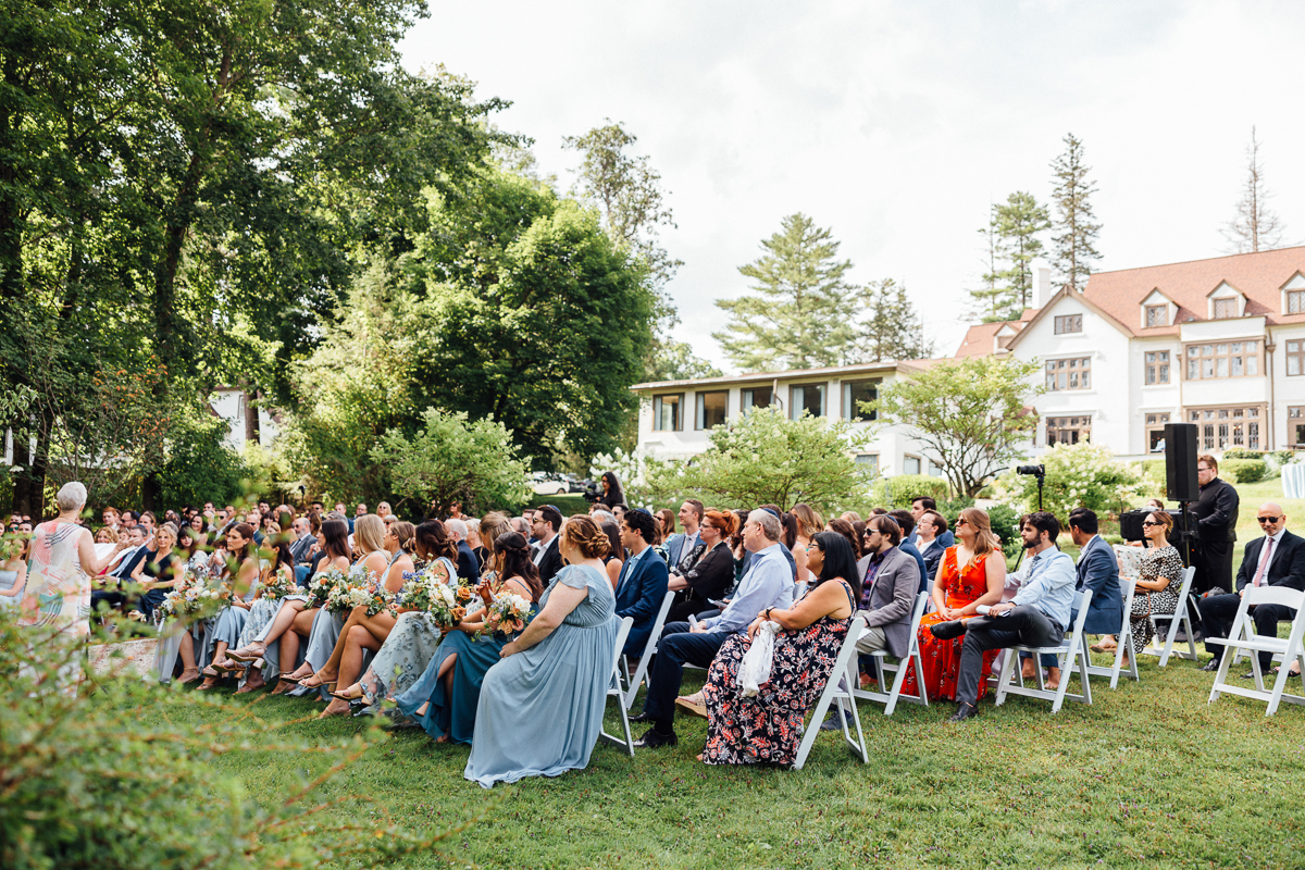 A large group of wedding guests sit on white chairs outdoors at Seven Hills Inn, surrounded by greenery, with a grand house in the background on a sunny Lenox Massachusetts wedding day. Everyone is dressed formally and facing the same direction.
