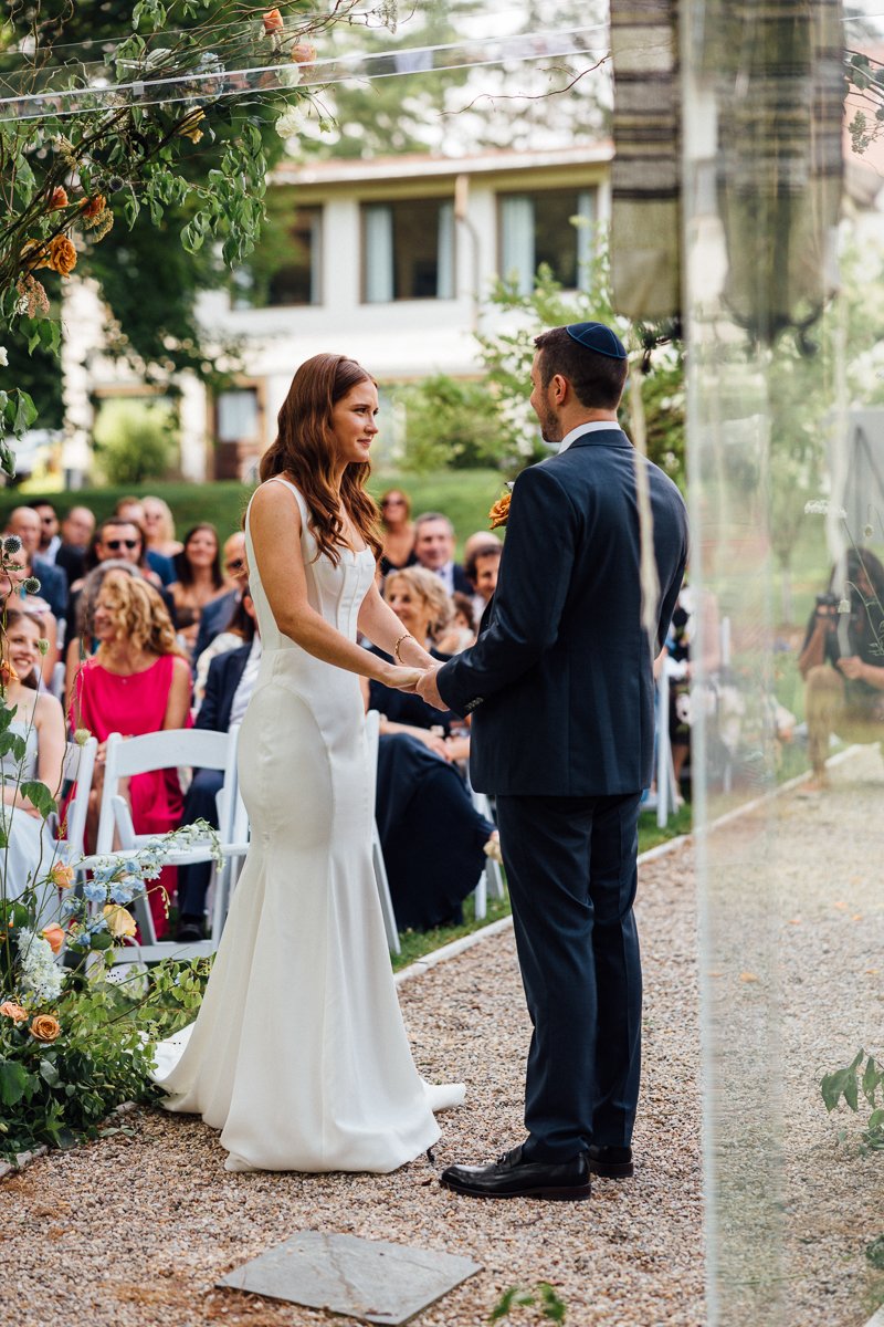 A bride and groom stand facing each other holding hands during an outdoor Lenox Wedding ceremony at Seven Hills Inn, with guests seated behind them and greenery surrounding the scene.