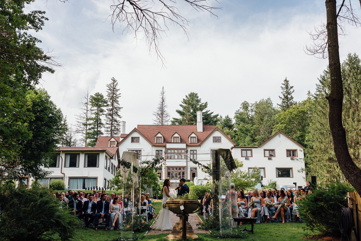 A bride and groom stand at an outdoor altar in front of a large white mansion at Seven Hills Inn, a stunning Lenox Massachusetts wedding venue, surrounded by guests on either side of a garden path with lush trees in the background.