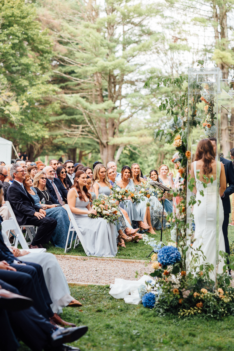 A bride in a white dress stands at an outdoor wedding ceremony at Seven Hills Inn in Lenox, MA, facing seated guests and bridesmaids in light blue dresses holding bouquets, surrounded by lush greenery and floral arrangements.
