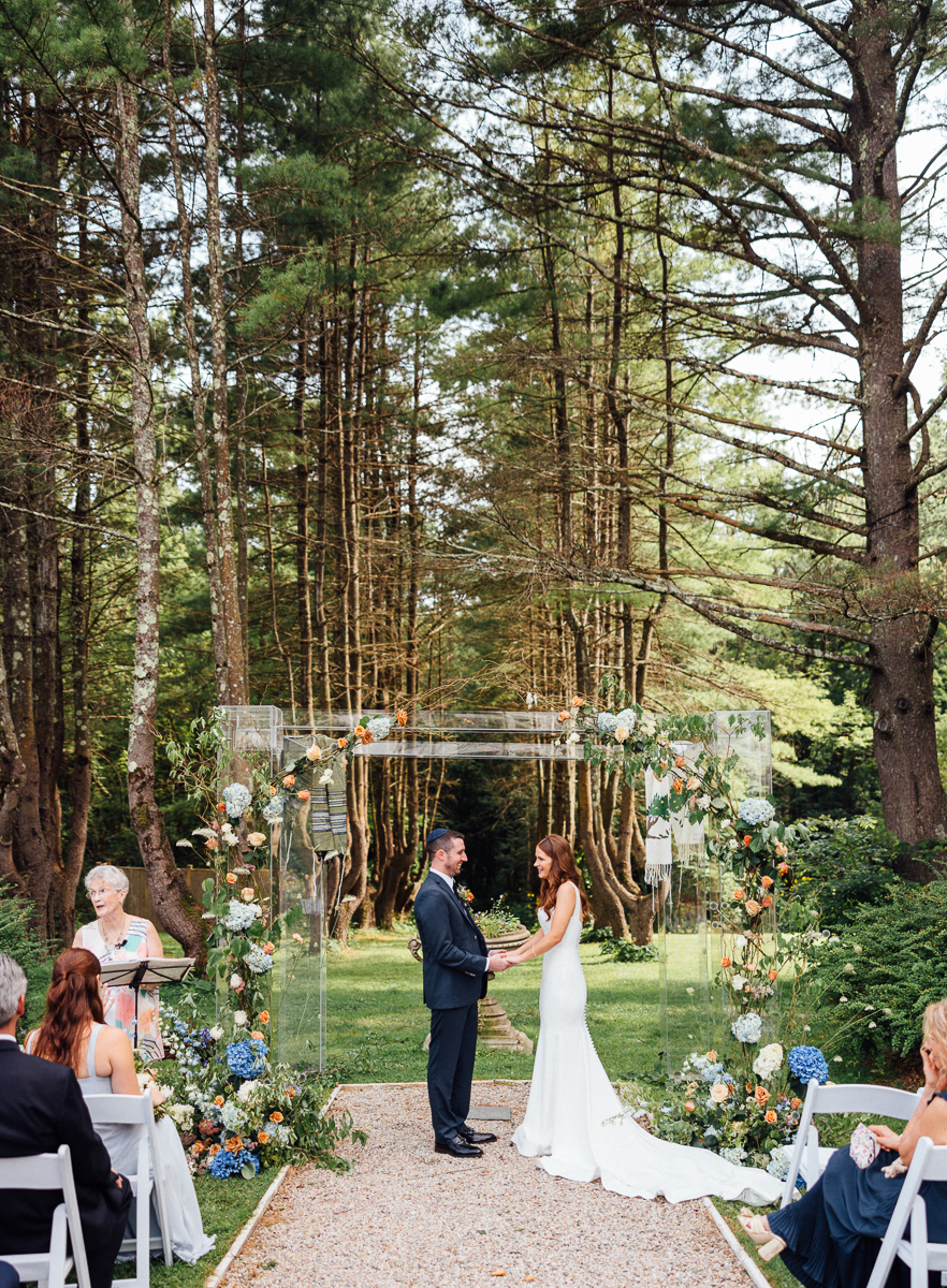 A bride and groom stand under a floral arch exchanging vows at their Seven Hills Inn wedding in Lenox, MA, surrounded by guests seated on white chairs, with towering trees framing the romantic outdoor ceremony.