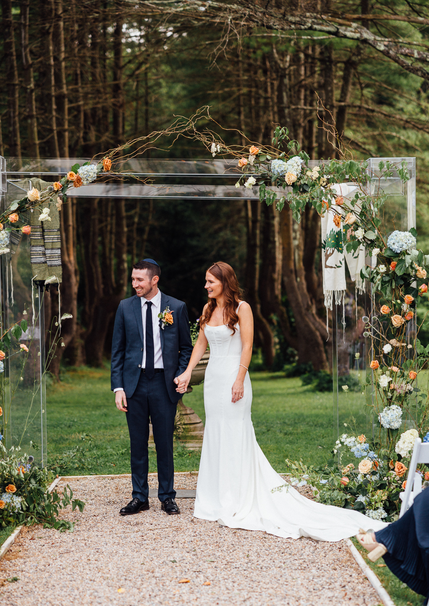 A bride in a white dress and a groom in a suit stand holding hands under a floral archway outdoors, surrounded by trees and greenery, during their Seven Hills Inn Wedding in Lenox MA.