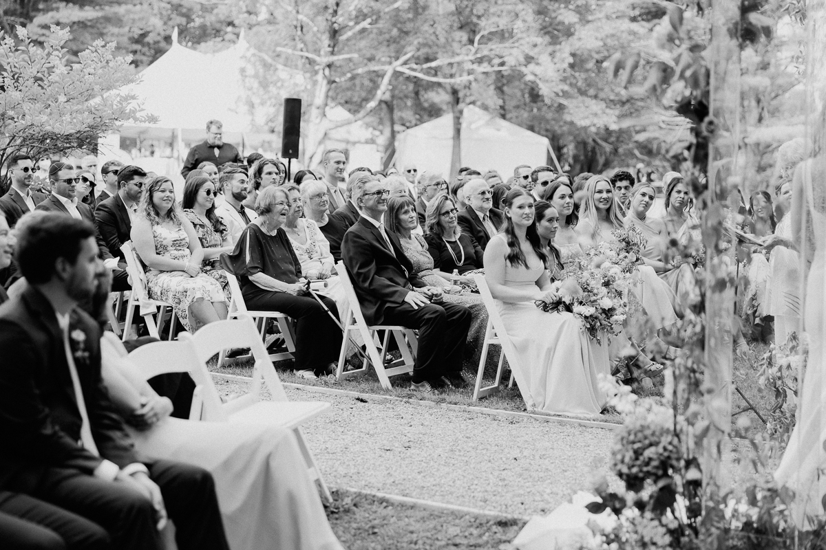 A black-and-white photo of wedding guests seated outdoors at a Seven Hills Inn Wedding in Lenox, MA, attentively watching the ceremony. Trees and tents form the backdrop, with bridesmaids holding bouquets seated in front.