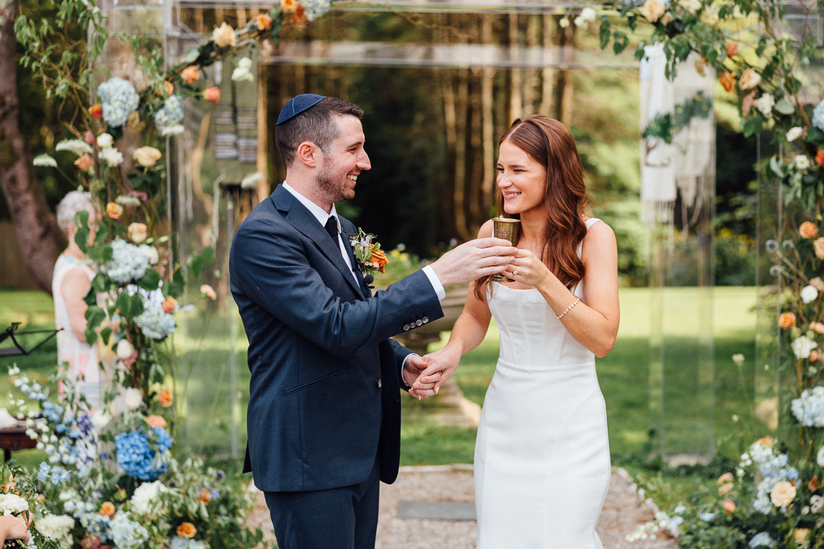 A bride and groom stand under a floral arch outdoors at their Seven Hills Inn wedding in Lenox, MA, smiling and holding a cup together. The groom wears a suit and yarmulke; the bride wears white, surrounded by lush greenery and flowers.