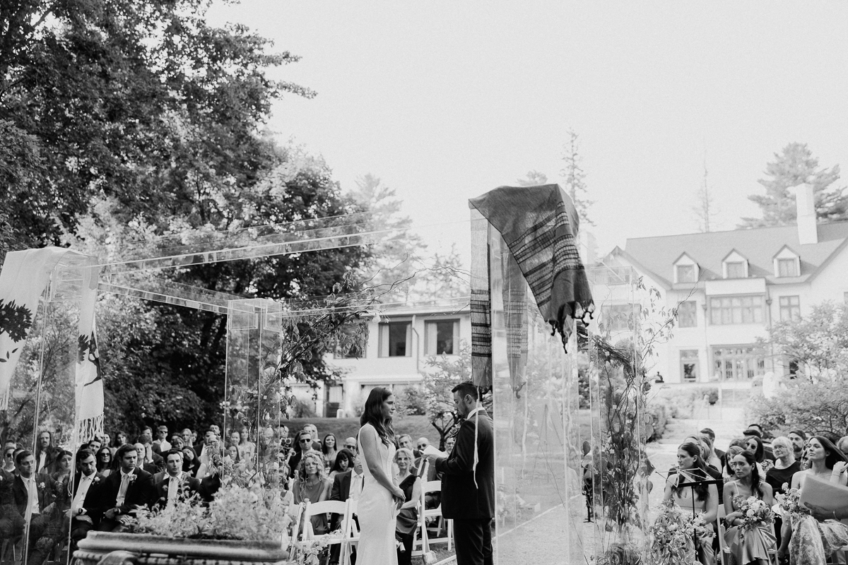 A bride and groom stand facing each other under a decorated wedding arch at their Seven Hills Inn wedding, surrounded by guests seated outdoors in Lenox MA. The scene is set in front of a large house, with trees and greenery around. The image is in black and white.