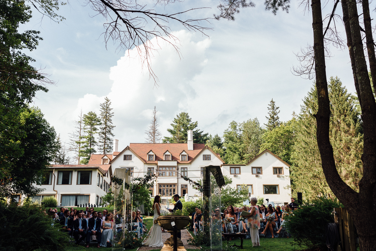 An outdoor wedding ceremony unfolds in front of a large white house surrounded by trees. Guests watch as the couple stands at the altar in the garden under a cloudy sky at this beautiful Seven Hills Inn wedding in Lenox, MA.