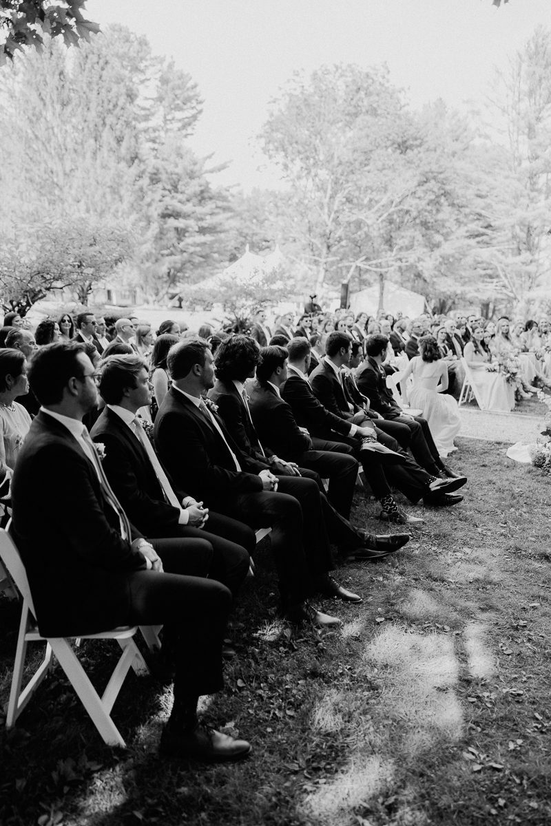 Black and white photo of people seated outdoors at a Lenox MA wedding ceremony, with men in suits on white chairs in the foreground and more guests and trees visible in the background.