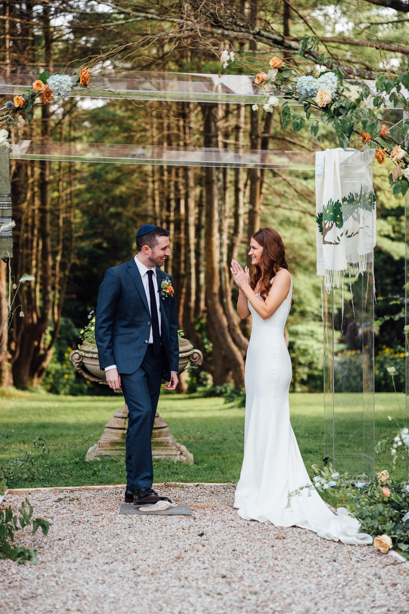 A bride in a white dress and a groom in a suit stand facing each other outdoors under a floral arch, surrounded by trees at their joyful Seven Hills Inn Wedding in Lenox MA, looking happy and emotional during the ceremony.
