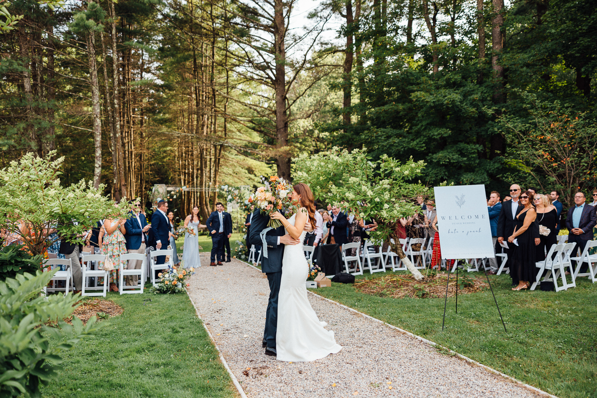 A bride and groom kiss at the end of the aisle during their Seven Hills Inn wedding in Lenox, MA. Guests seated on white chairs among lush green trees look on, while a welcome sign stands on the right side of the path.