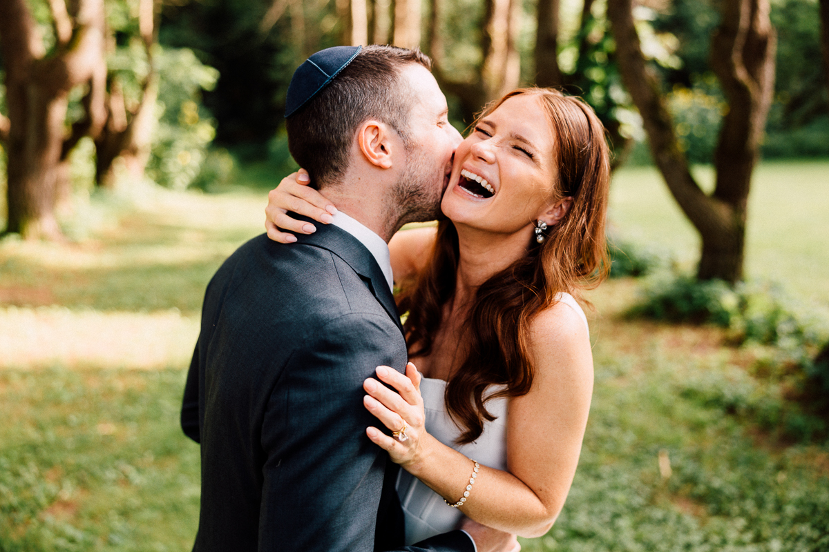 A smiling couple embraces outdoors at their Seven Hills Inn wedding in Lenox, MA; the man in a suit and kippah playfully kisses the laughing woman in a white dress, framed by lush trees and greenery.