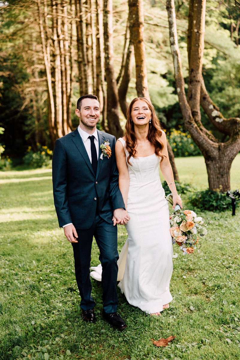 A smiling bride and groom walk hand in hand on the grass at Seven Hills Inn in Lenox, MA, surrounded by trees. The bride wears a white dress and holds a bouquet, while the groom wears a dark suit with a boutonnière for their wedding.