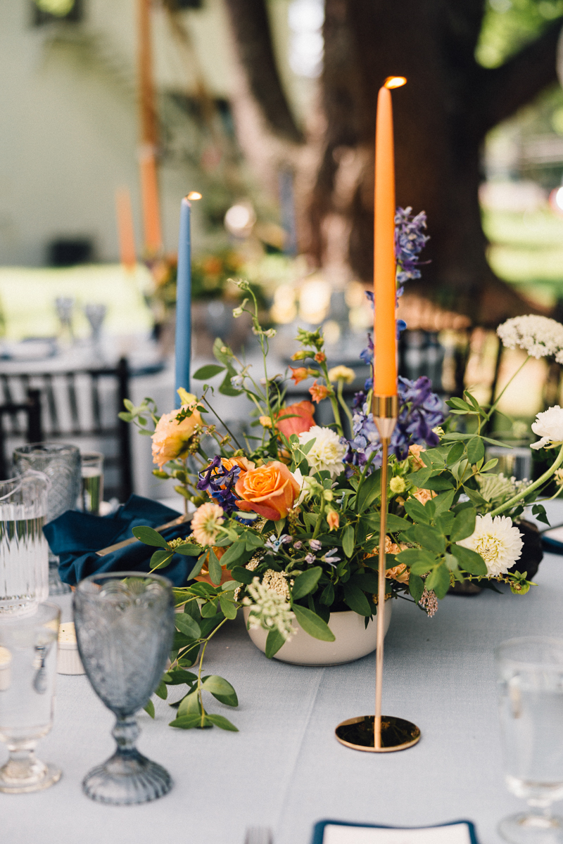 A table set outdoors at a Lenox MA Seven Hills Inn wedding features elegant glassware, blue napkins, and a floral centerpiece. Tall orange and blue taper candles add a colorful, sophisticated touch to this charming wedding setting.