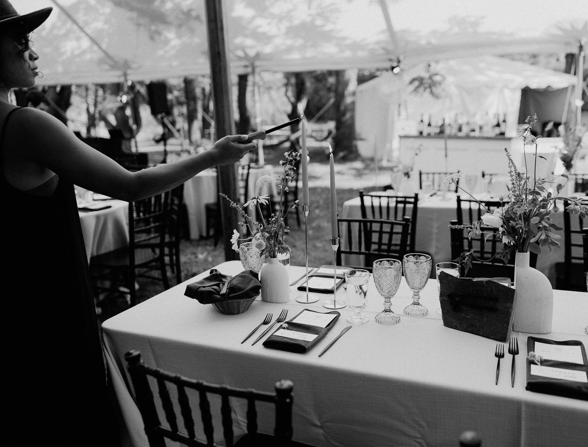 A woman in a hat arranges items on an elegantly set outdoor table with glassware, plates, candles, and floral centerpieces; black and white photo from a Seven Hills Inn wedding in Lenox MA, with empty chairs visible.