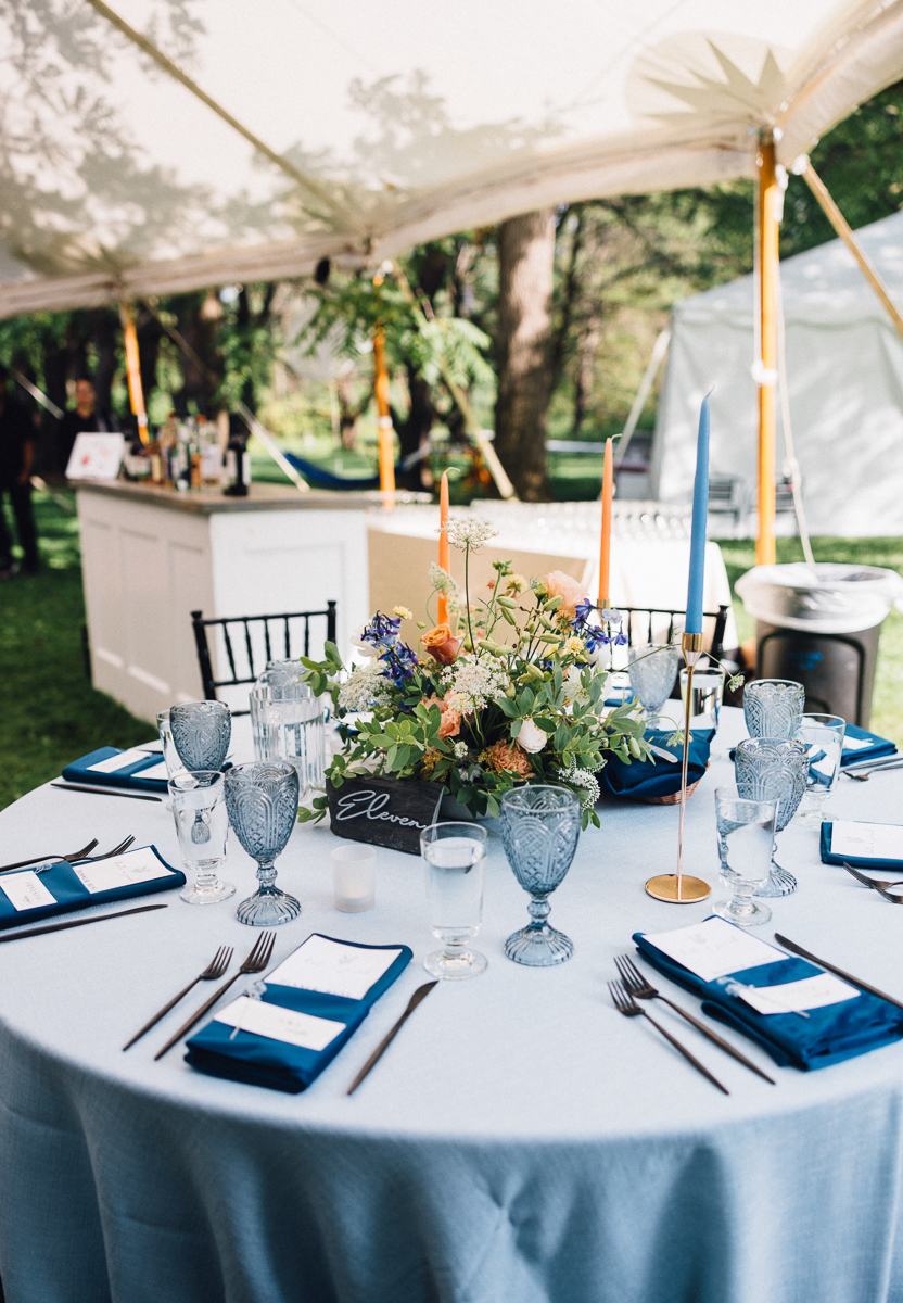 A round table set for a Seven Hills Inn wedding in Lenox MA features blue napkins, clear glassware, a colorful floral centerpiece, tall candles, and a table number sign. A bar and lush greenery are visible in the background under a tent.