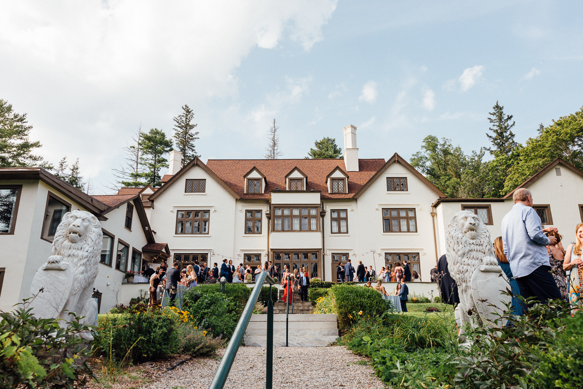 A large group of people gathers outside the grand, white Seven Hills Inn mansion with a red roof in Lenox, MA on a sunny day. Stone lion statues flank the entrance, and lush greenery surrounds this picturesque wedding scene.